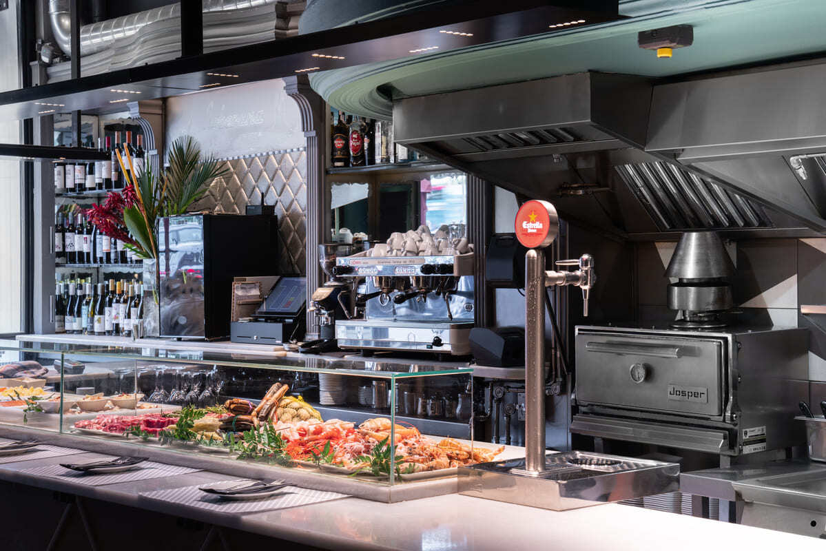 View over counter at Vinitus, Barcelona. Bar area has variety of proteins and vegetables under glass. Behind on the back wall is a cash register, bottles of wine and Josper charcoal oven. 